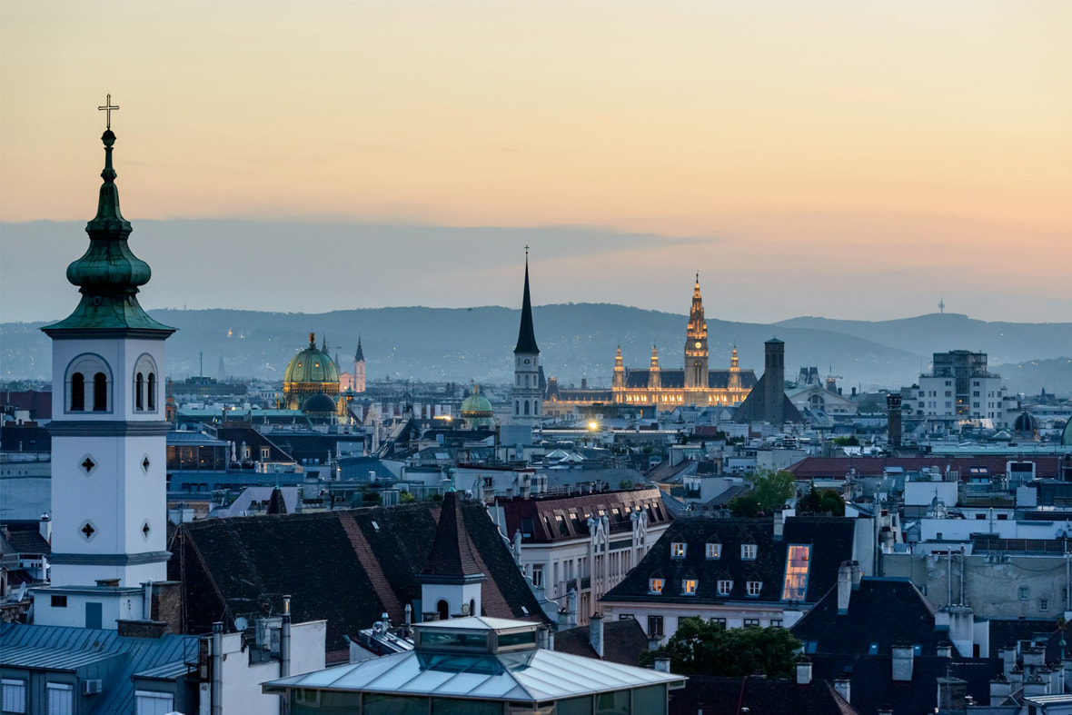 Blick über Wien bei Sonnenuntergang mit Kirchtürmen und dem beleuchteten Wiener Rathaus im Hintergrund.