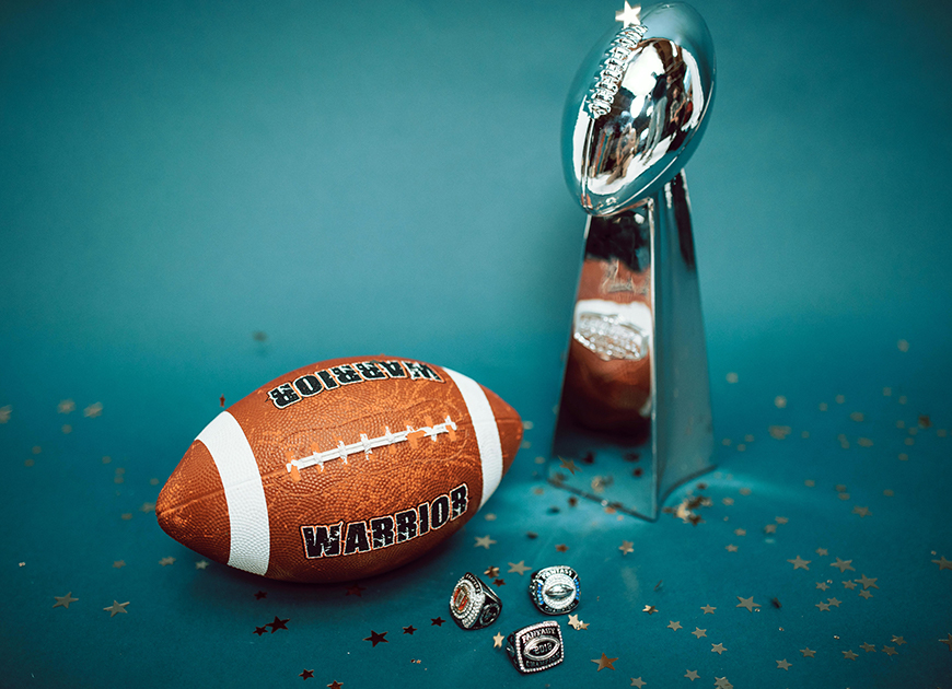 Football mit der Aufschrift "Warriors" und eine silberne Trophäe und drei NFL-Meisterschaftsringe auf einem blauen Hintergrund.